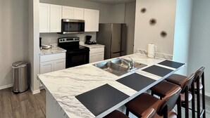 Una cocina con armarios blancos, electrodomésticos de acero inoxidable, encimera de mármol y una zona de bar con taburetes.