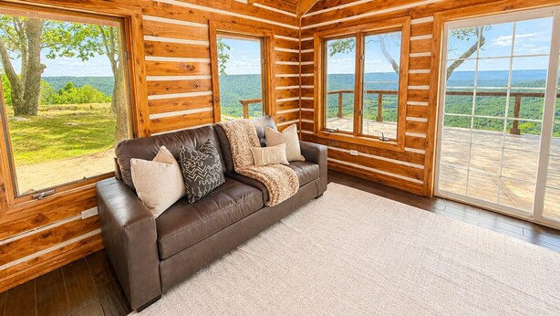 A log cabin interior with a leather couch, large windows, and a view of a forested landscape. - One Bedroom Cabin with Panoramic View at Paul's Point (Altamont)