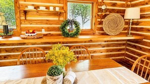 A wooden cabin interior with a dining table, chairs, a lamp, a shelf with dishes, and a window with a view of trees. - One Bedroom Cabin with Panoramic View at Paul's Point (Altamont)