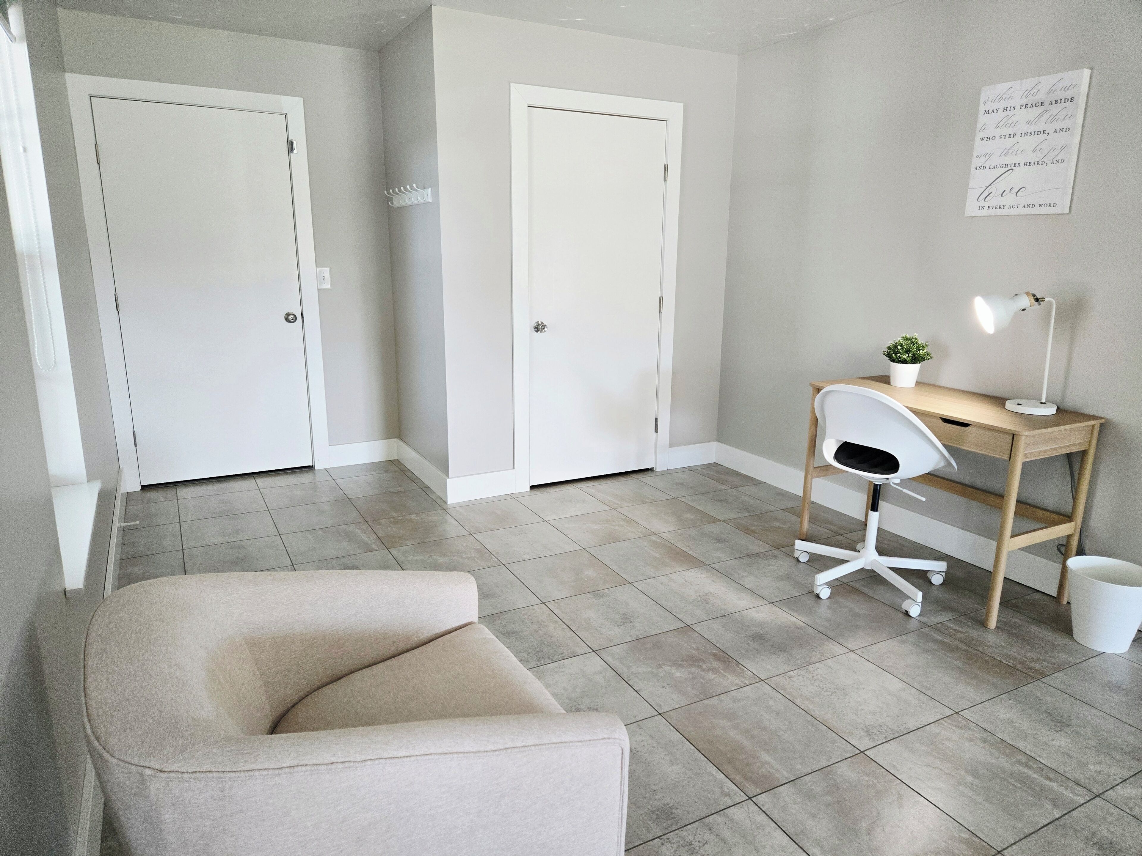 Une pièce comprenant un fauteuil beige, un bureau en bois avec une chaise, un lampadaire blanc et une petite plante en pot.