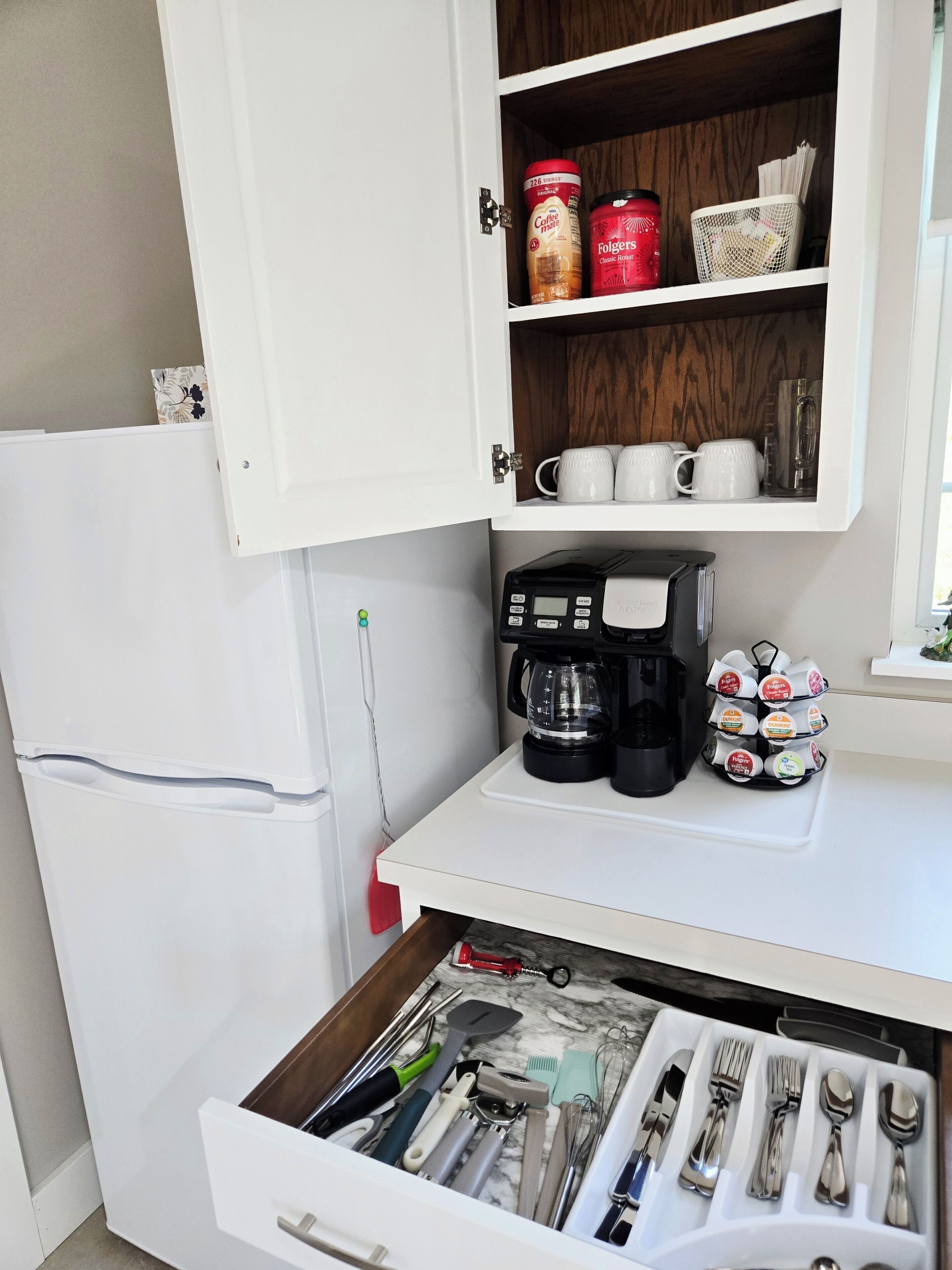 Una cocina con un refrigerador blanco, una cafetera, un escurreplatos y un cajón con utensilios.