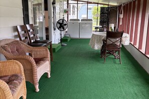 Une pièce meublée de mobilier en osier, avec un ventilateur, une table recouverte d’une nappe à motifs floraux et une vue sur l’extérieur.