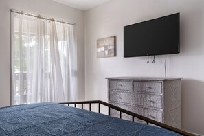 A bedroom with a bed, a television mounted on the wall, a window with sheer curtains, and a woven cabinet.