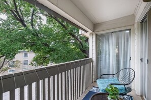 A balcony with a chair and a plant, a door with curtains, and a view of trees and buildings.