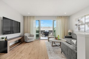 A modern living room with a flat-screen TV, a sofa, a coffee table, and a sliding glass door leading to an outdoor area. - Sunlit Slate, 3BR+Bonus+XBOX+BBQ with Modern Design + Private Deck (Calgary)
