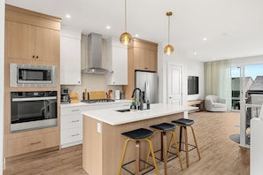 A modern kitchen with a central island, bar stools, and a view of the outdoors. - Sunlit Slate, 3BR+Bonus+XBOX+BBQ with Modern Design + Private Deck (Calgary)