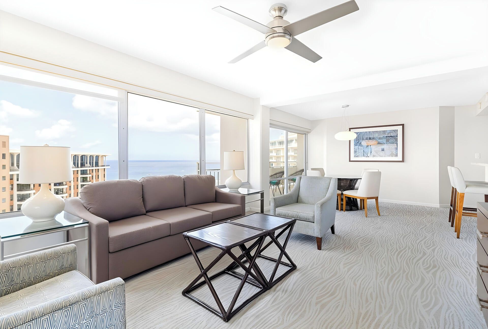 A modern living room with a sofa, coffee table, and chairs, large windows with a view, and a ceiling fan.