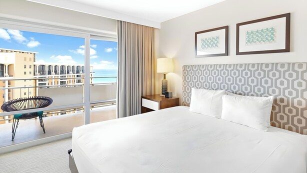 Quarto de hotel com uma cama grande, uma escrivaninha com luminária, uma cadeira e vista para o mar através de uma varanda.