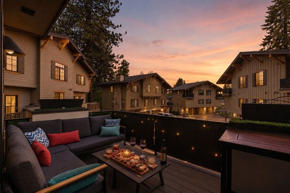A deck with a sofa, table, and snacks, overlooking a row of buildings at sunset. - Downtown McCall Retreat | Walk Everywhere (McCall)