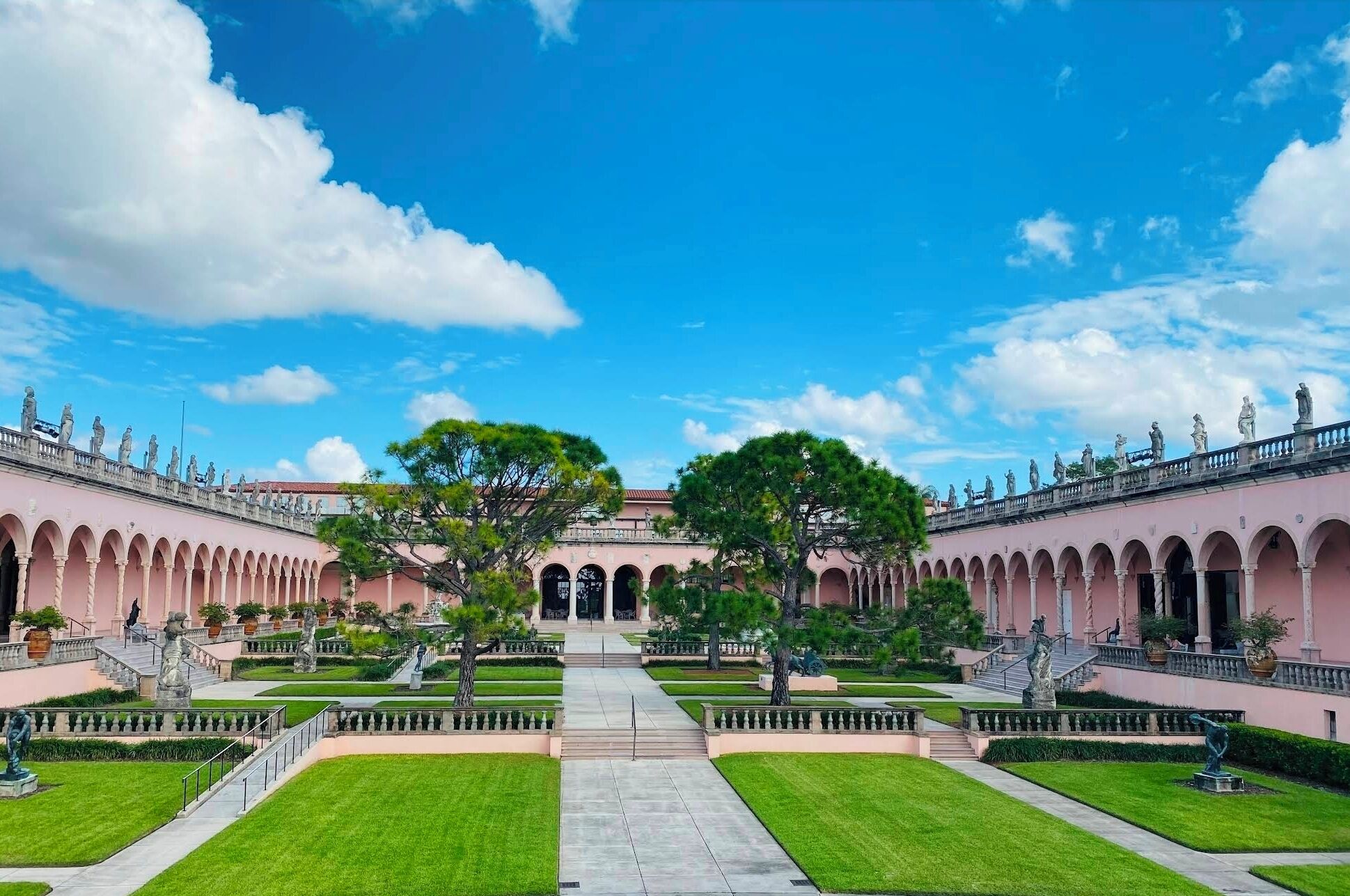 A symmetrical courtyard with a central pathway, flanked by pink buildings with arched windows and statues.
