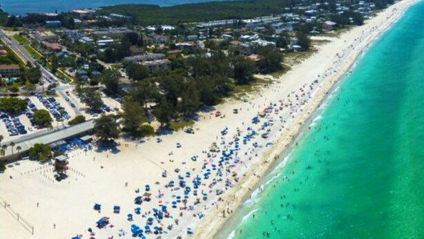 Vista aérea de uma praia com mar azul-turquesa cristalino, areal arenoso e área residencial densa com casas e carros estacionados.