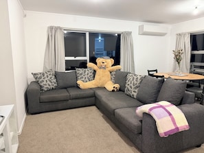 A living room with a sectional sofa, a teddy bear, a dining table, and a vase with flowers. - 3BR Home - Walk to Train, Lynn Mall & Westgate (Auckland)