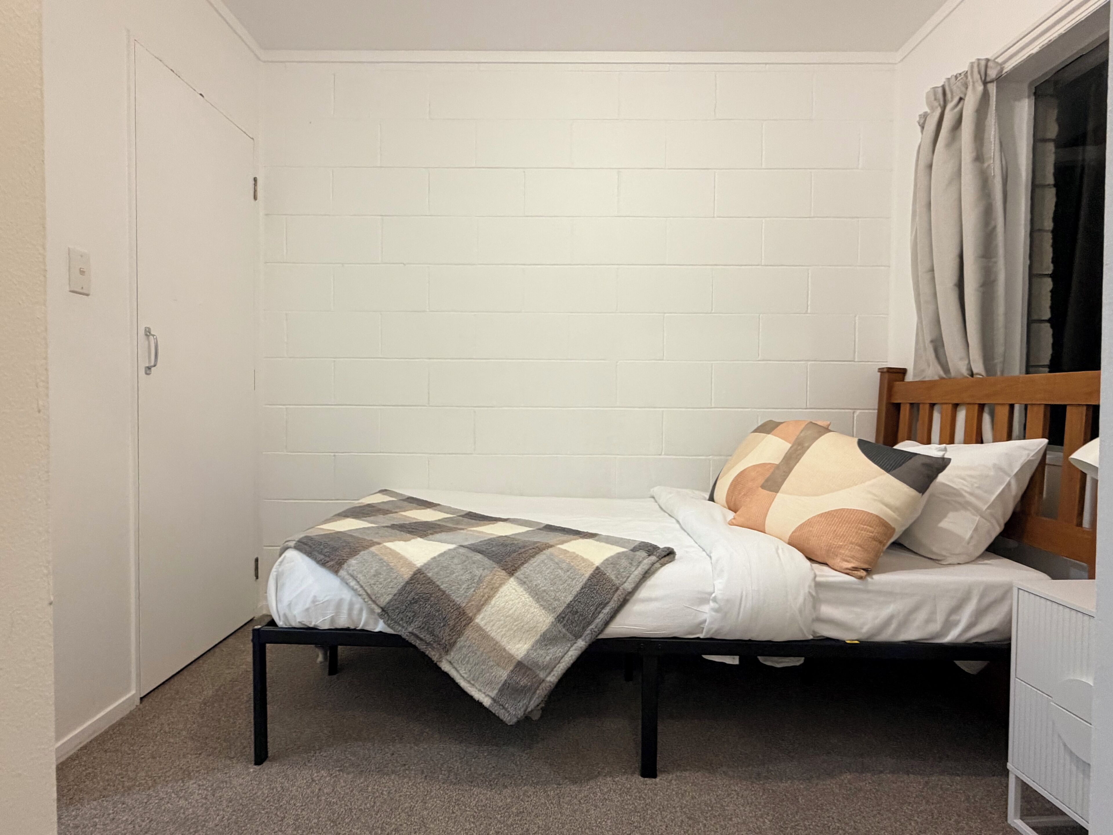 Un lit simple avec une couverture à carreaux, deux oreillers, une tête de lit en bois et une armoire blanche.