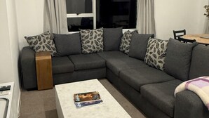 En stue med en grå hjørnesofa, et hvidt sofabord og et vindue med gardiner.