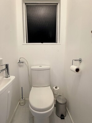 Et lille, hvidt badeværelse med toilet, vask, et vindue og en skraldespand.