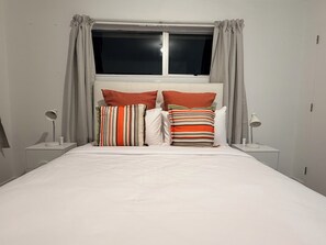 Uma cama com roupa de cama branca, duas mesas de cabeceira com luminárias e uma janela com cortinas.