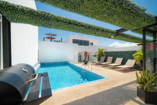 Ein Swimmingpool mit Liegestühlen, einem Grill und einem modernen Gebäude mit weißer Fassade.