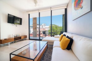 A modern living room with a flat-screen TV, a sofa with yellow and black cushions, a glass coffee table, and a view of the outdoors through large windows.