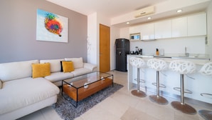 A modern living room with a sofa, coffee table, and a kitchen area with white cabinets and bar stools. - Apartment with Balcony & Pool (Playa del Carmen)