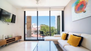 A modern living room with a glass coffee table, a white sofa with yellow cushions, a mounted TV, and a large window offering a view of the outdoors. - Apartment with Balcony & Pool (Playa del Carmen)