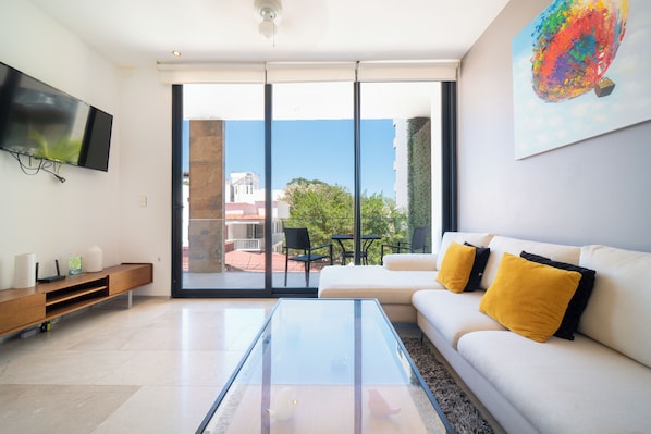 A modern living room with a glass coffee table, a white sofa with yellow cushions, a mounted TV, and a large window offering a view of the outdoors.