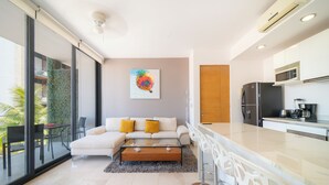 A modern living room with a glass partition, a sofa, a coffee table, and a kitchen area with a refrigerator and microwave. - Apartment with Balcony & Pool (Playa del Carmen)