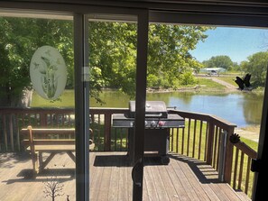 Une terrasse avec un barbecue, un banc et une vue sur un lac bordé d’arbres, avec un véhicule visible au loin.