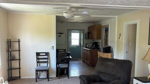 Un salon avec un canapé, une table basse, un fauteuil, une bibliothèque et, au fond, un coin cuisine.