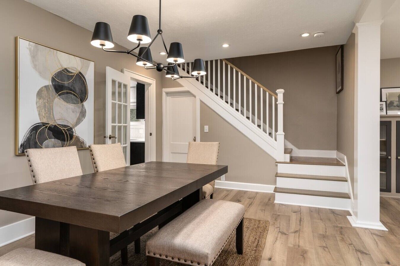 Een eetruimte met een donker houten tafel, beige fauteuils, een moderne kroonluchter en een trap met witte leuningen.