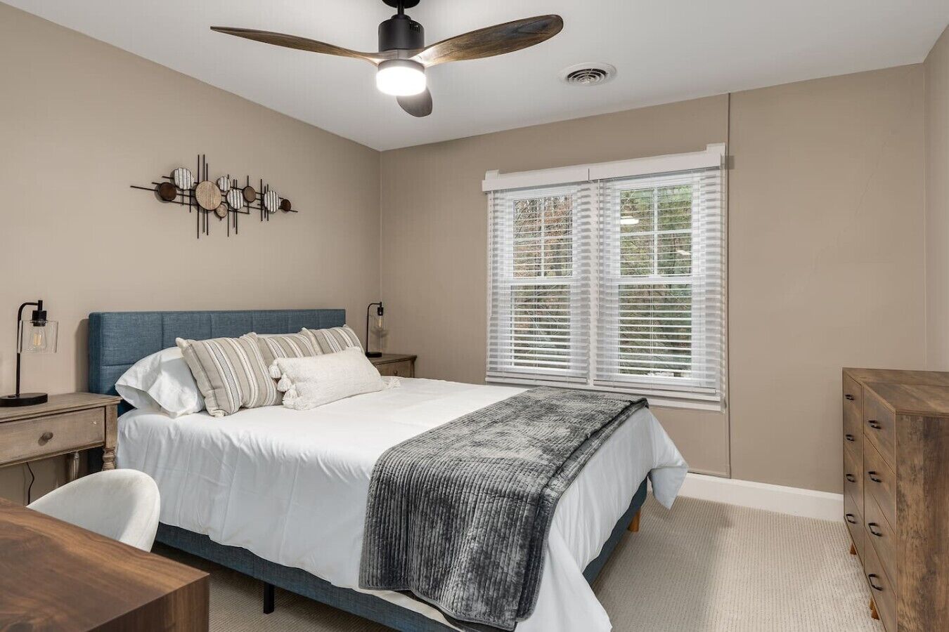 Een slaapkamer met een bed, een bureau, een stoel, een dressoir, een plafondventilator en een raam met jaloezieën.