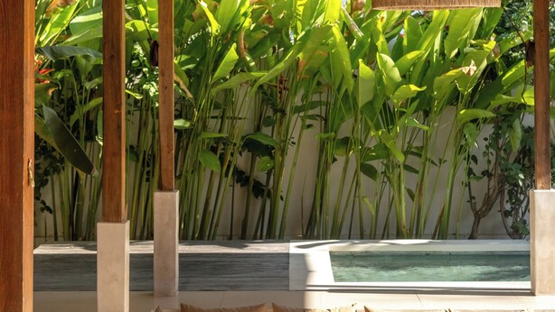 Quarto com telhado de colmo, amplo espaço de assentos acolchoados, luminária pendente e vista para uma piscina e vegetação exuberante.