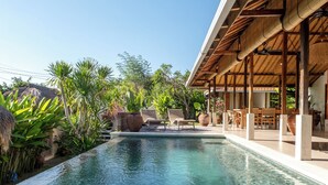 Piscina com área coberta, cercada por plantas tropicais e céu claro.