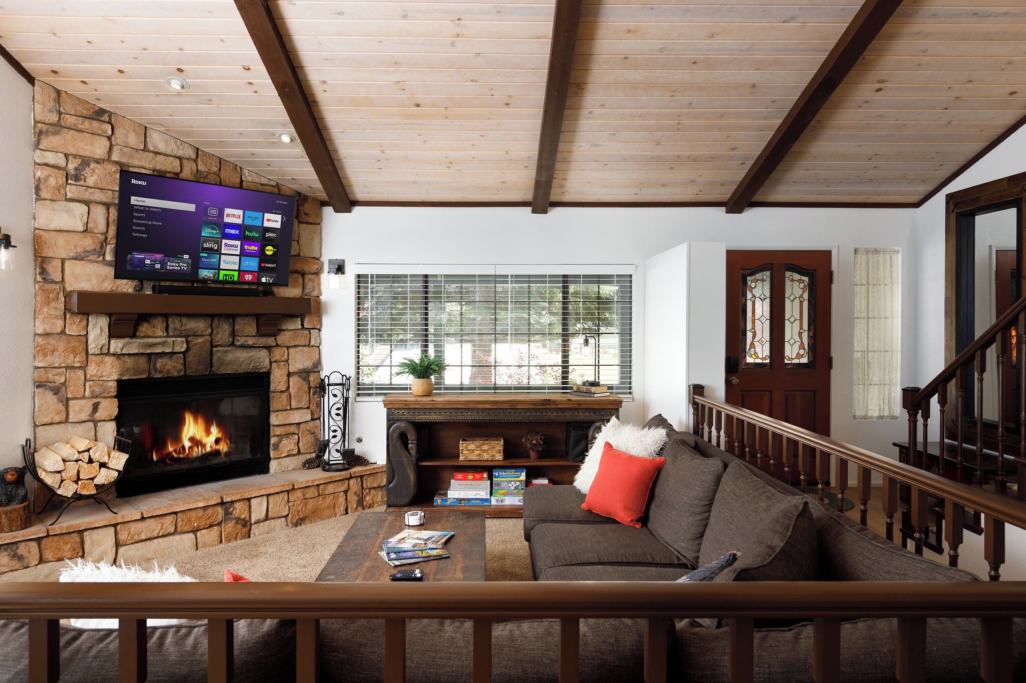 A living room with a stone fireplace, a flat-screen TV mounted above it, a sofa with a red pillow, and a staircase with wooden railings.