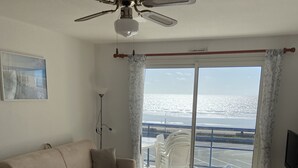 Een woonkamer met een beige bank, een plafondventilator, een televisie en uitzicht op het strand door een groot raam.