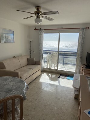 Ein Wohnzimmer mit beiger Couch, Deckenventilator, Fernseher und Blick auf den Strand durch ein großes Fenster.