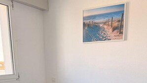 Een net opgemaakt bed met blauw beddengoed, een bedlampje en een schilderij van een strandgezicht aan de muur.