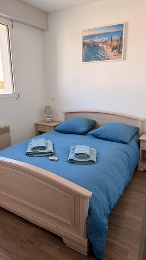 Ein ordentlich bezogenes Bett mit blauer Bettwäsche, eine Nachttischlampe und ein Bild einer Strandansicht an der Wand.