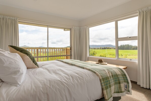 A bedroom with a large bed, a wooden headboard, a nightstand, and a view of a green field through large windows.