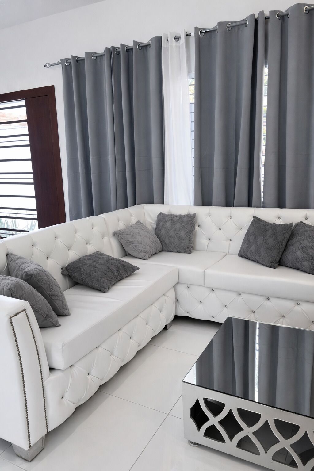 Um sofá branco modular com almofadas cinzas, uma mesa de centro de vidro, cortinas cinzas e uma parede branca.