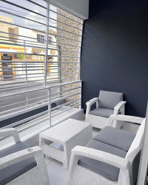 Un balcon agrémenté de meubles en osier peints en blanc, d’un fauteuil rembourré de couleur grise et d’un mur en pierre.