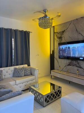 A modern living room with a sofa, a glass coffee table, a TV mounted on a marble wall, and a ceiling fan.