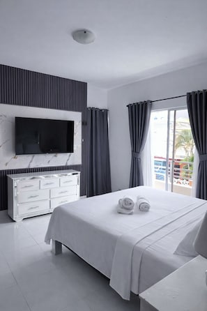 A modern hotel room with a bed, a flat-screen TV mounted on a marble wall, a white dresser, and a balcony with curtains.