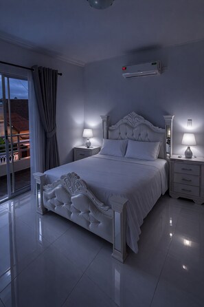 A bedroom with a large bed, two bedside tables, a ceiling-mounted air conditioning unit, and a view through a glass door.