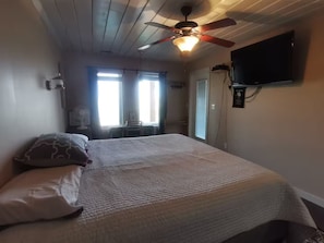 A bedroom with a bed, ceiling fan, TV, and a window with curtains.