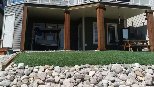 Ein zweistöckiges Haus mit Balkon, überdachter Veranda und einer Steinmauer zur Geländeanhebung.