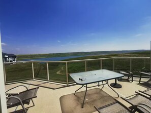 A balcony with a glass table and chairs, overlooking a lake and green landscape.