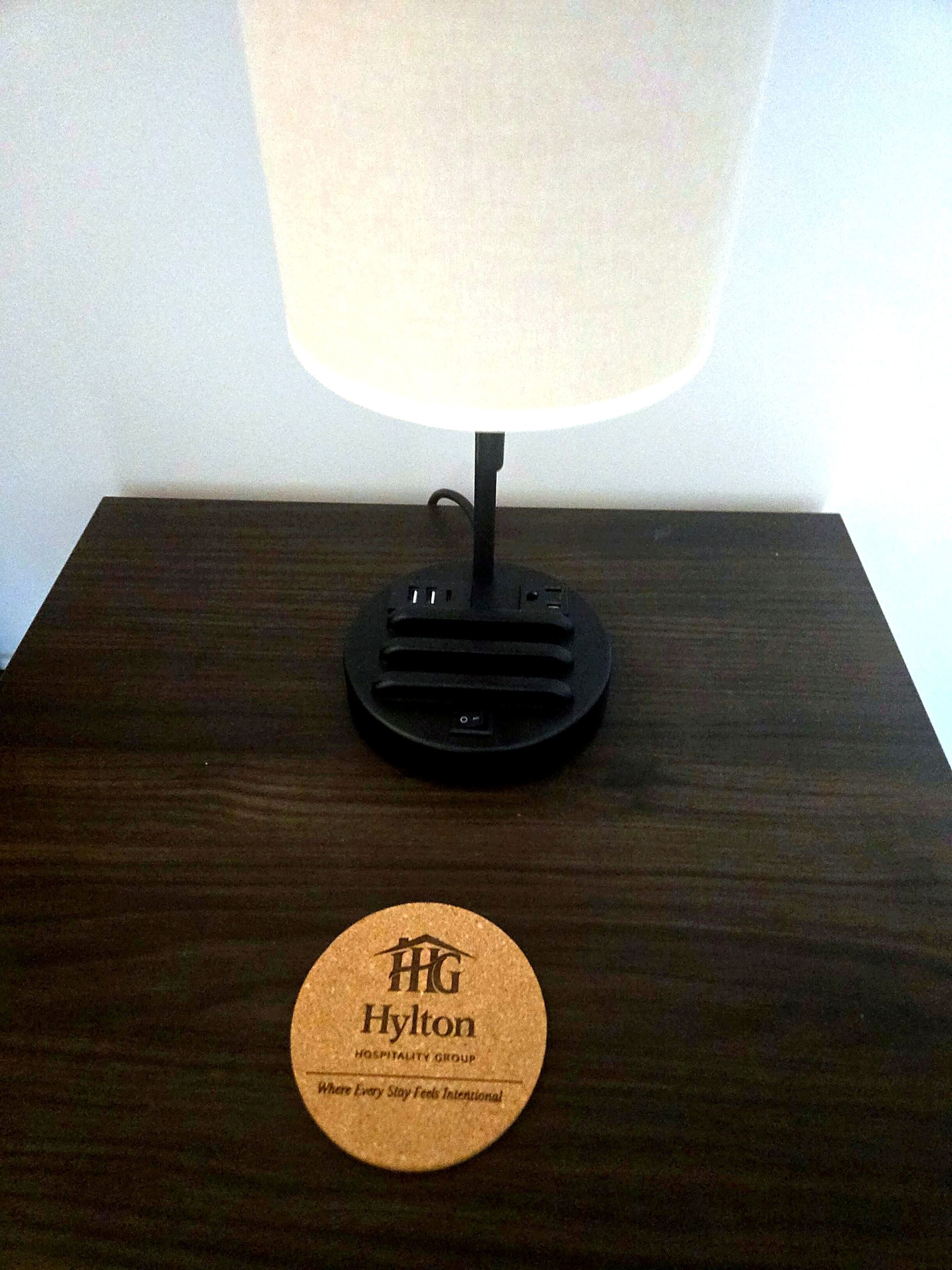 A wooden nightstand with a black lamp and a circular coaster with 'HG Hylton Hospitality Group' text.