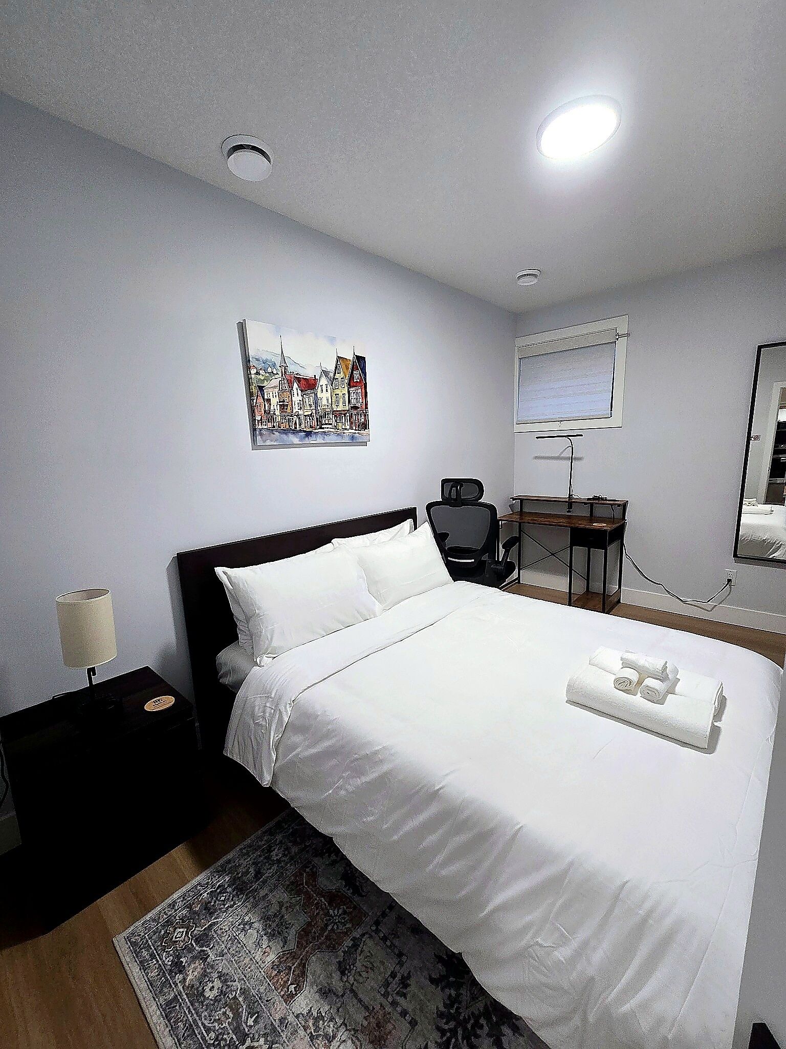 A hotel room with a bed, a desk, a chair, a lamp, and a painting on the wall.