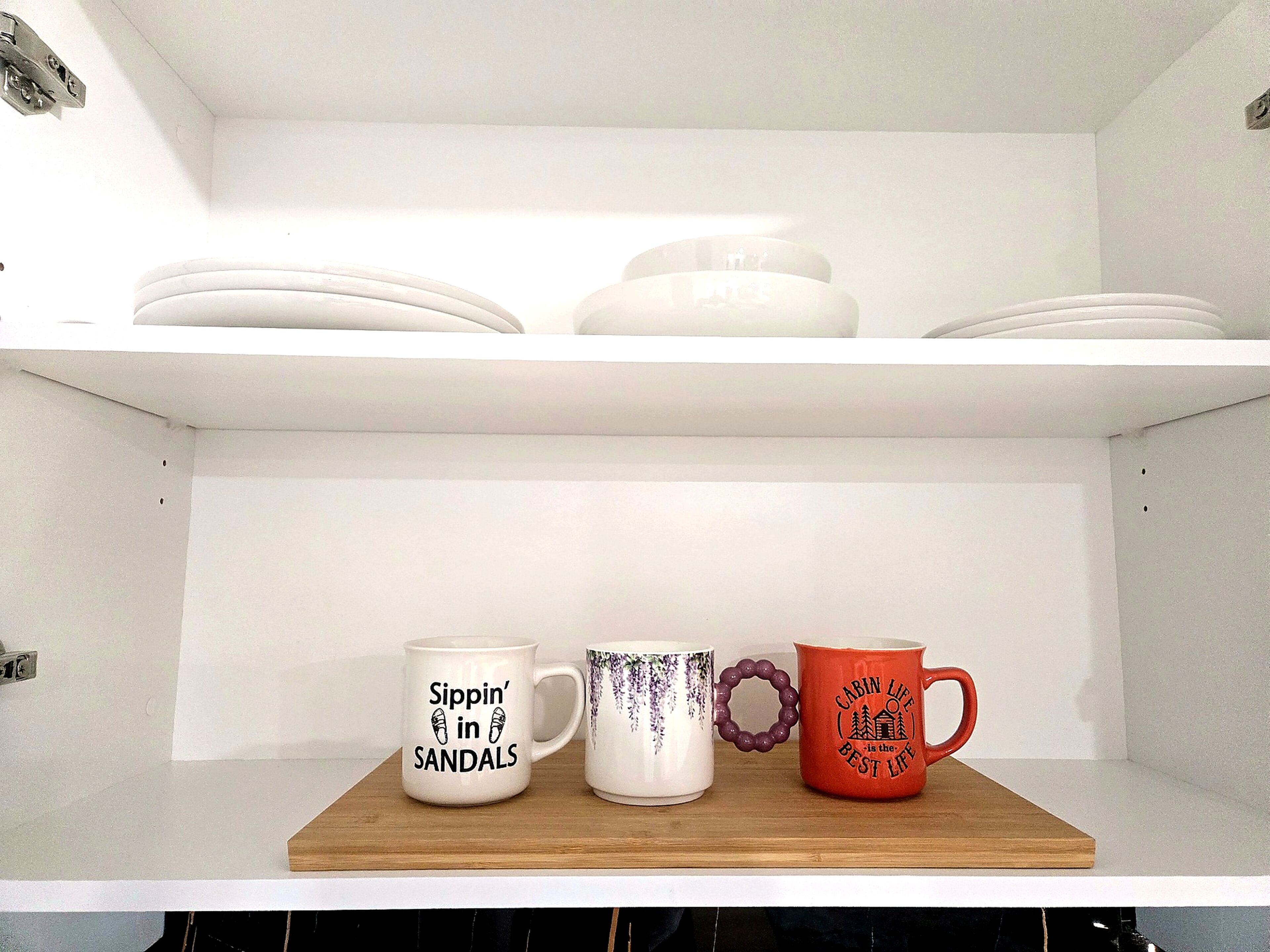 A shelf with white plates, a white mug with black text, a white mug with purple patterns, and an orange mug with black text.
