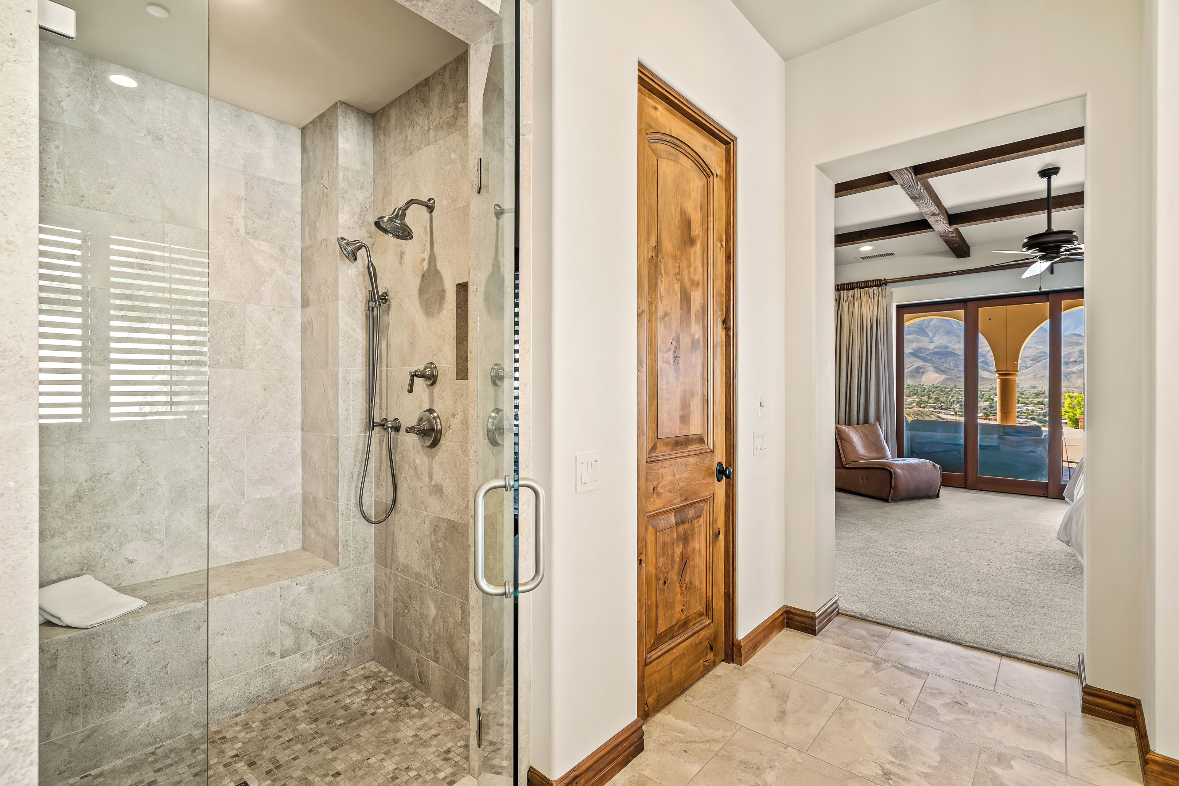 A modern bathroom with a glass-enclosed shower, a wooden door, and a view to a balcony with outdoor seating.